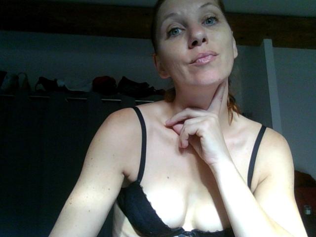 ApoloniaRed with pouty lips, blue eyes, and brown hair, wearing black lace bra, touching chin, showing small tits and fair skin, in dimly lit room.