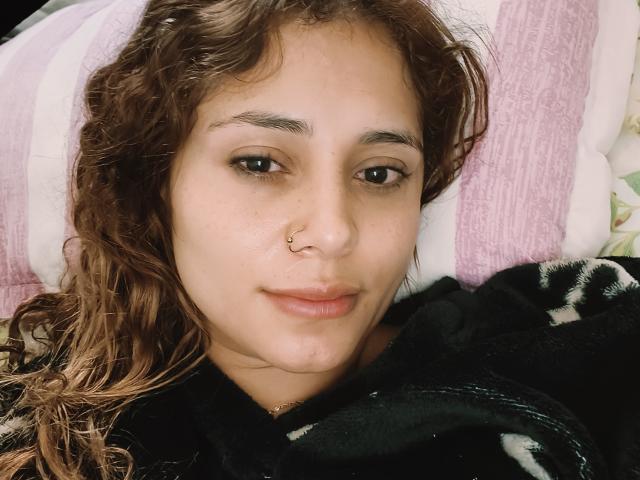 EliBaker with curly brown hair, nose ring, subtle smile, and slightly flushed cheeks, lying on a pink and white pillow, wearing a black fleece.