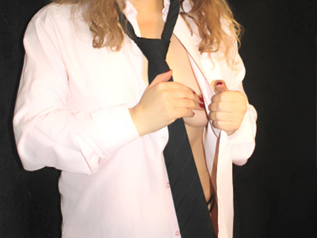 Priscilla, een volwassen vrouw met krullend blond haar, draagt een roze shirt en een zwarte stropdas. Ze houdt haar shirt open om haar blote borsten te laten zien, waarbij haar tepels zichtbaar zijn. Zwarte achtergrond..