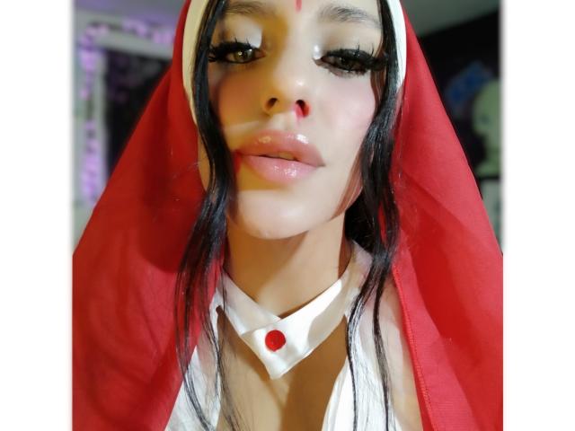 ErotiicAlexa with black hair, wearing a red nun's veil and white collar, has glossy lips, a red dot on her forehead, and a white choker with a red gem.