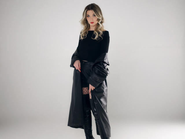InvitingTina with blonde curls, black long-sleeve top, black leather coat, black shorts, fishnet stockings, black boots, standing, holding coat, red lipstick, sexy, confident.
