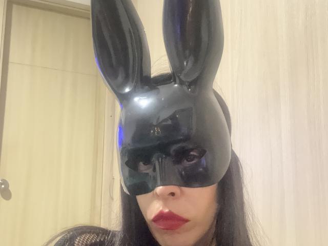 SeleneBrigth wearing a shiny black bunny mask with tall ears, red lipstick, and long black hair, with a mesh top, in a bright room.