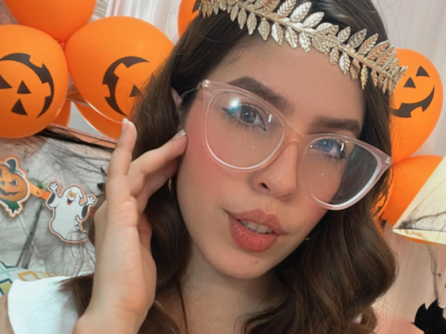 NatyBakers mit vollen, rosafarbenen Lippen, klaren Brillen, braunen, gewellten Haaren, einem goldenen Kopfschmuck und orangefarbenen Ballons im Hintergrund. Hände, die das Gesicht berühren. Kürbisgesichter auf den Ballons..