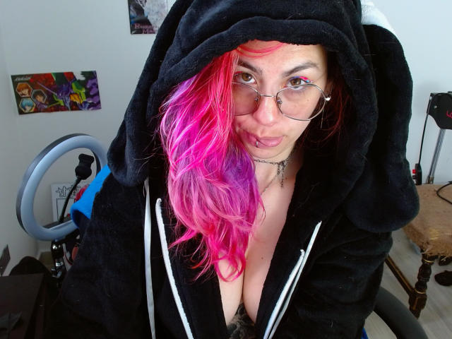 NatashaLau with pink and red hair, glasses, black hoodie with ears, showing cleavage, slightly open hoodie, and a nose ring, sitting in front of a camera ring light.