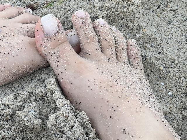 Destinis' dirty, sandy feet with toes covered in white pebbles, showing slight tan skin and small, scattered black grains, resting on a sandy surface.