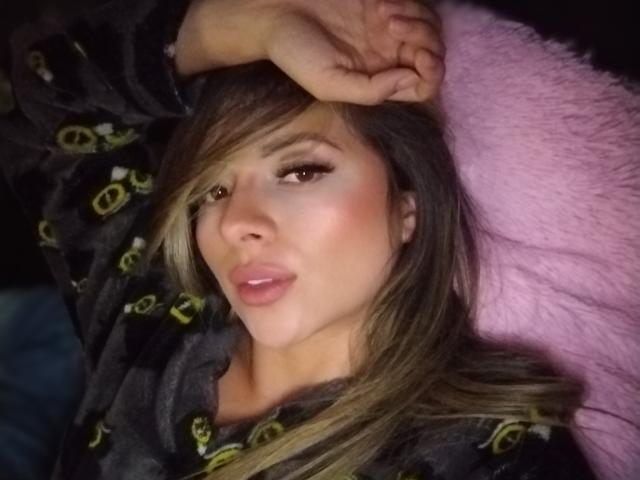 LaraHottie with pouty lips, lying down, wearing black pajamas with green logos, right hand on forehead, long blonde hair, pink pillow, subtle makeup, and a cute, sexy face.