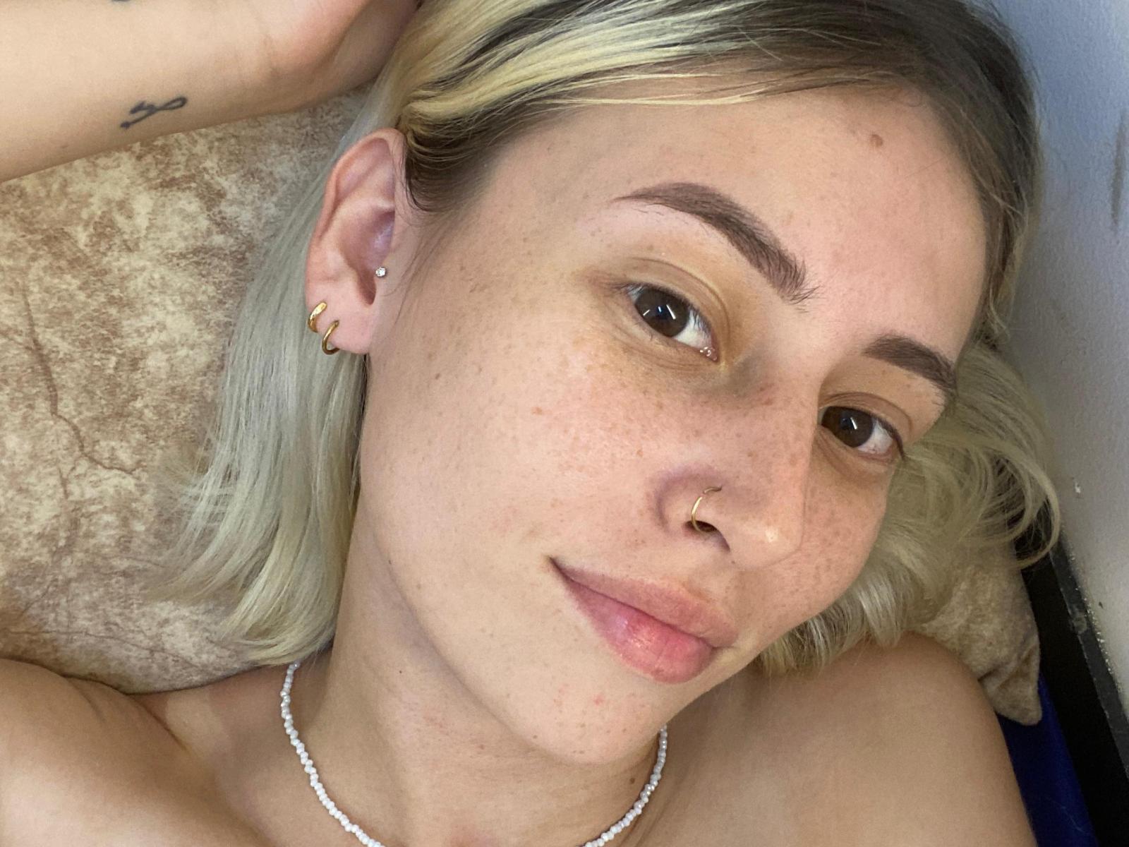 SelenaAmour tem cabelo loiro, olhos castanhos, sardas, um piercing no nariz, pequenas argolas de ouro nos ouvidos, um colar de pérolas e uma tatuagem no braço direito. Ela está deitada em um travesseiro..