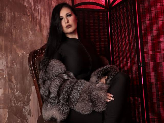 Kelly Kent, atractiva, con cabello oscuro y ondulado, vestida con una blusa negra con cremallera y una chaqueta de piel gris esponjosa, sentada en una silla de madera, luciendo seductora con iluminación roja..