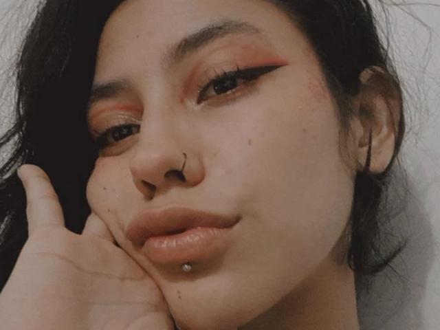 MiaUtena with dark hair, red eyeshadow, black eyeliner, nose ring, lip piercing, and a slight smile, touching her cheek. Her skin is light with small freckles.