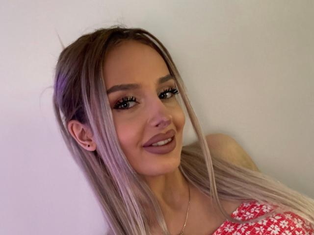 RheaI has long, straight, blonde hair, brown eyebrows, and a smile with dark lipstick. She's wearing a red floral top and has a gold necklace.