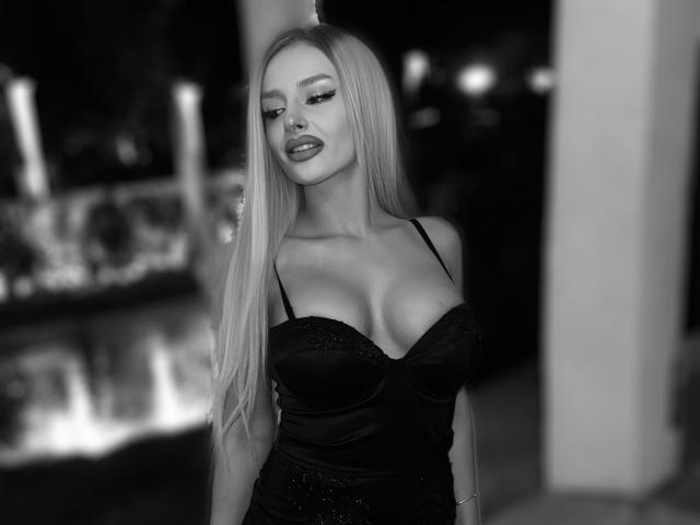RheaI has long blonde hair, large boobs in a black, sparkly dress with spaghetti straps, and a confident, sexy expression in a black-and-white photo.