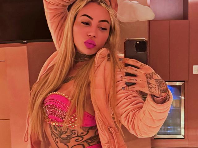 TatuadaSafadaHot with long blonde hair, pink lipstick, and tattoos, wearing a pink strapless top and pink jacket, taking a mirror selfie, showing her large, shiny tits.