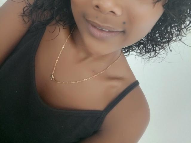 Lunah69 with dark curly hair, black tank top, gold chain necklace, brown skin, small black beauty mark on upper lip, smiling slightly, showing collarbone and shoulder.