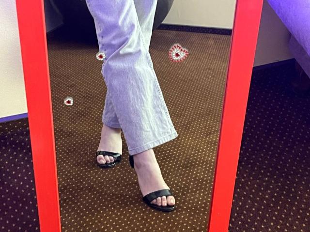 Las piernas de Valissia, vestida con jeans azul claro y sandalias negras de punta abierta, están frente a un espejo con marco rojo, sobre una alfombra marrón con pequeños puntos blancos, y hay una flor roja en el suelo..