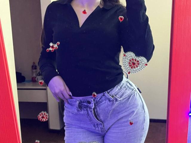 Valissia, met haar ronde heupen, draagt een zwart shirt met een V-hals en lichtblauwe, hoge taille jeans. Haar hand zit in haar zak, en op haar shirt en jeans staan kleine, rode hartjes..