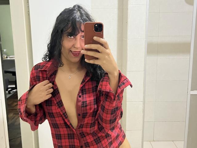 QueenMel, com cabelos pretos cacheados, vestindo uma camisa xadrez vermelha desabotoada, expondo seus seios, tirando uma selfie no espelho com um celular marrom, sorrindo e usando um colar..