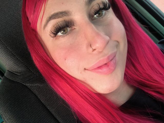 SammyViolet with bright pink hair, green eyes, long black eyelashes, nose ring, and tongue slightly out, sitting in a car, looking at camera with a cute smile.