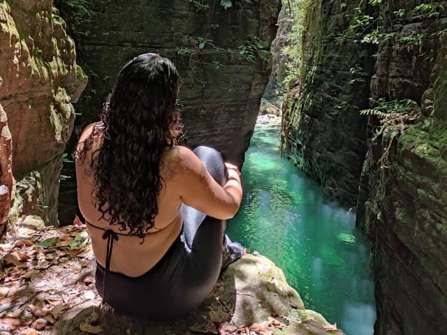 QueenMel, con i capelli neri ricci, di spalle alla telecamera, seduta su delle rocce, indossa un top da bikini nero e pantaloni neri, e guarda un canyon roccioso verde, pieno di acqua turchese..