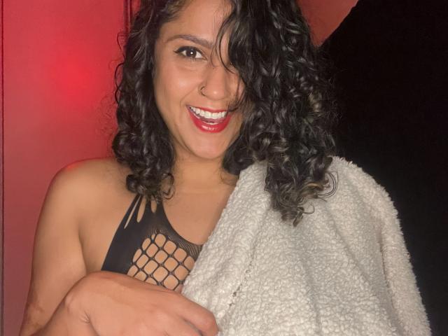 QueenMel, com cabelo preto encaracolado, piercing no nariz, batom vermelho, sorrindo, vestindo uma blusa de rede preta, um cobertor branco felpudo, segurando os seios, luz vermelha ao fundo..