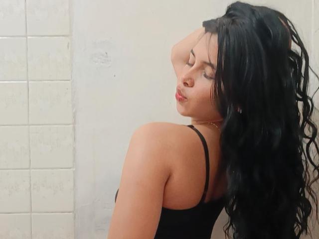 MistressAsshley, avec ses cheveux noirs ondulés, les yeux fermés, portant un débardeur noir, sa main dans ses cheveux, montrant son épaule nue et une légère vue de ses fesses..