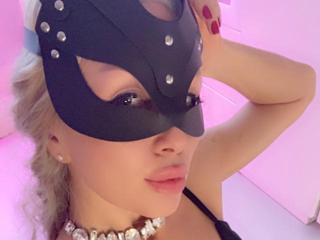 AvaGreenx with a black mask, red nails, pearl necklace, and platinum blonde hair, showing off her tits and a sexy face. Pink lighting in the background.
