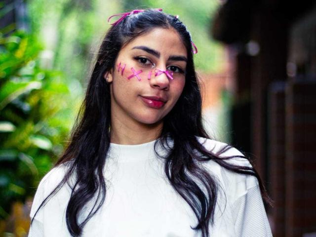 JessicaSoler has long dark hair, pink butterfly tattoos on her face, and a white shirt, standing with a slight smile, looking cute and attractive.
