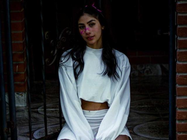 JessicaSoler with long dark hair, pink face paint, wearing a white long-sleeve crop top and matching white shorts, sitting between brick walls and black metal bars.