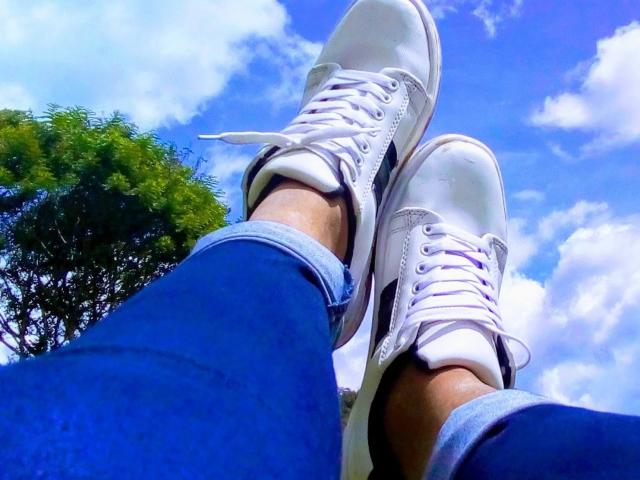 SophieSonyas' legs in white sneakers, blue jeans, and a sunny blue sky with fluffy clouds. Her feet are crossed, showing white laces and a tree to the left.