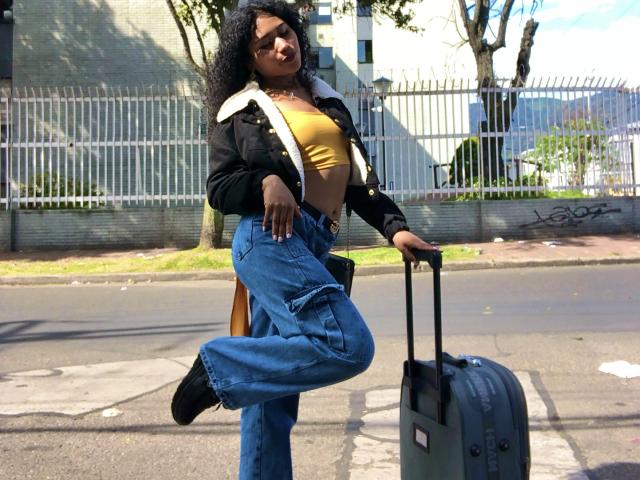 SophieSonyas with curly black hair, yellow top, black jacket, blue jeans, and black boots, standing on street, holding gray suitcase, one leg raised, looking down.