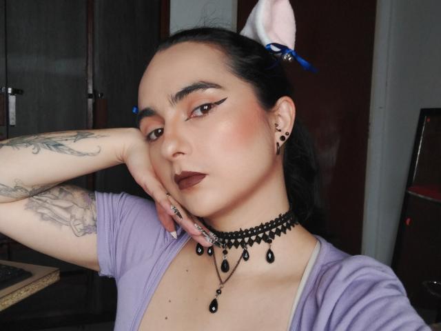 CandyParis a un maquillage sombre, un collier noir, un haut violet, des tatouages sur le bras, des yeux marron, un rouge à lèvres foncé, des pinces à cheveux bleues et des boucles d'oreilles noires. Elle pose avec une main sur la joue..