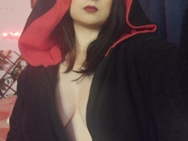 CandyParis with black and red hooded robe, showing deep cleavage, pale skin, dark lipstick, and straight black hair. Her tits are partially visible through the open robe.