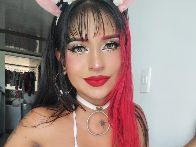 Nefertiti69x with red-black hair, cat ears, white choker, red lipstick, and blue eyes, showing off her tits and a black star tattoo near her neck.