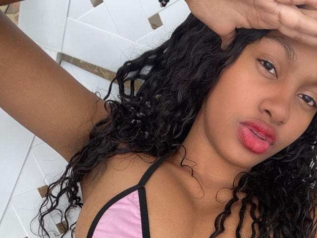 StormiHills with curly black hair, pink-and-black bikini top, and red lips, covering her forehead with one hand, showing her medium brown skin and small tits.