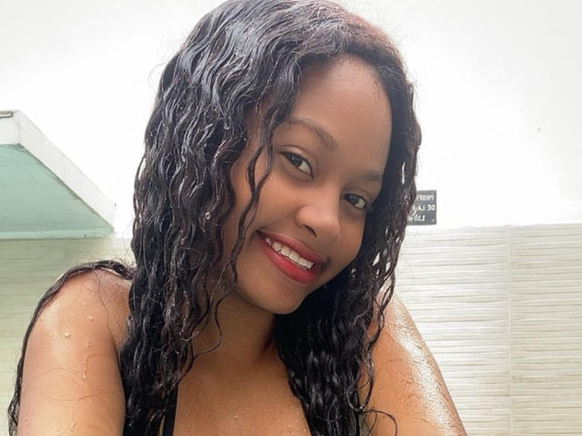 StormiHills with wet, curly black hair, smiling with red lipstick, showing her tits, glistening with water in a bright, white bathroom.