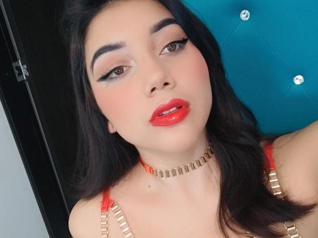 SquirtinGamy with red lipstick, black eyebrows, and long dark hair, wearing a gold choker and red bra straps, looking at the camera with a slightly open mouth.