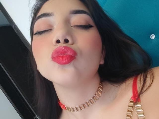 SquirtinGamy has her eyes closed, pouting with bright red lipstick, wearing a gold chain necklace and red bra straps, with long dark hair and thick eyebrows.