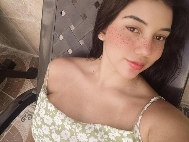 SquirtinGamy has freckled cheeks, pink lips, and brown eyes, wearing a green floral dress, with dark hair, sitting on a woven chair, showing her boobs.