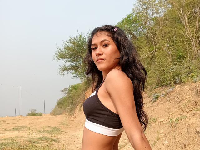 AnnieDarling with long black hair, wearing a black and white sports bra, looking over her shoulder with a slight smile, standing on a dirt path with trees.