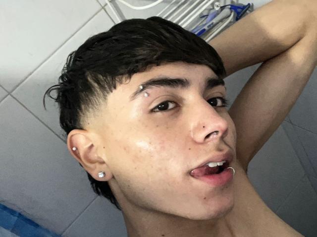 ImJuanda with short black hair, earrings, nose and facial piercings, tongue out, wet skin, looking at camera, arm raised. Shower background with blue bottle.