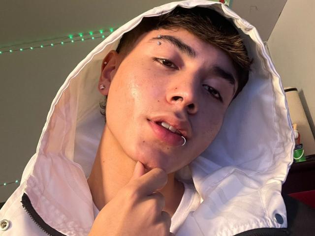ImJuanda with a nose piercing, lip ring, and ear studs, wearing a white hooded jacket, touching their chin, with short brown hair, and green string lights in the background.