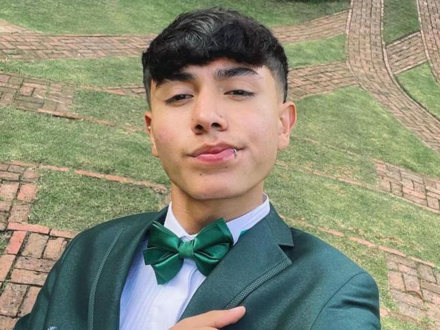 ImJuanda with dark hair, green bowtie, green jacket, and white shirt, sticking out pink tongue, holding jacket, cute, medium skin tone, standing on grass with brick paths.
