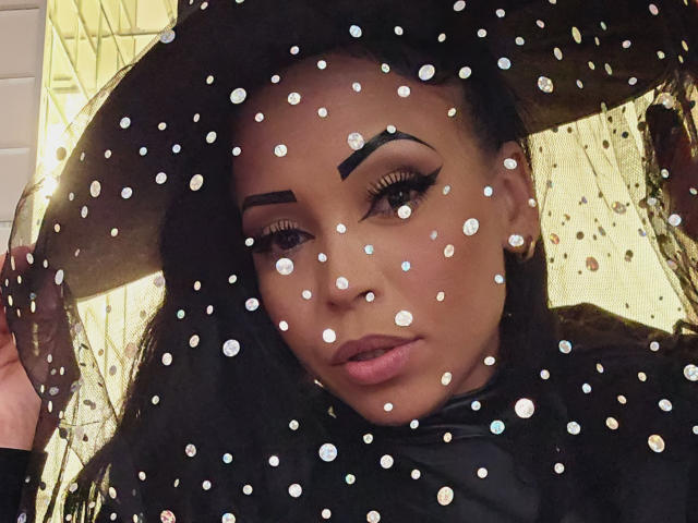 SquirtLatinQueen with dark skin, thick eyebrows, long eyelashes, and full pink lips, wearing a black hat, and gold hoop earrings, with white polka dots on a transparent surface.