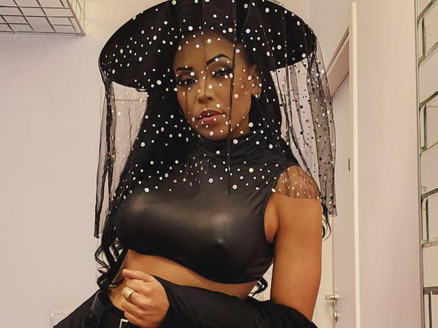 SquirtLatinQueen with dark skin, long black hair, wearing a sheer black polka-dot hat and black leather crop top, exposing her tits, holding a black garment in a white room.