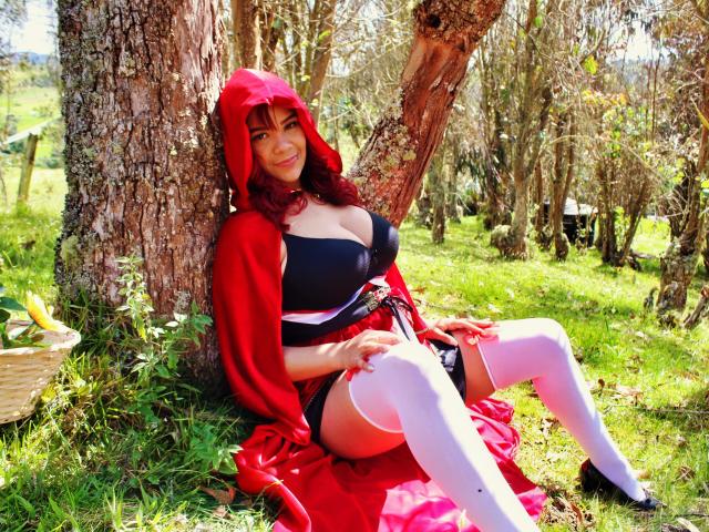 VictoriaField with red hood, black bra, white thigh-highs, and red cape, sitting against tree, smiling, showing her big tits, red panties, and white garter.