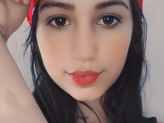 MileenaLily's face, with dark eyebrows, big brown eyes, red lipstick, and black hair, holding a red headband, showing a slight smile, and a small nose piercing.