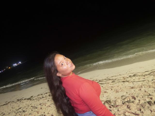 ElChicletv with long dark hair, red long-sleeve shirt, and blue jeans, looking back at the camera, showing her tits, on a sandy beach at night.