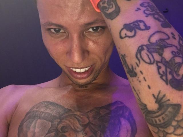 ElChicletv with a bald head, tattoos on arms and chest, smiling with white teeth, holding up a hand, visible chest and arm tattoos, purple lighting.
