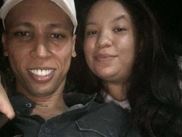 ElChicletv with dark skin and white cap, smiling, showing teeth, next to ElChicletv with light skin and long black hair, both wearing denim, in a dimly lit selfie.