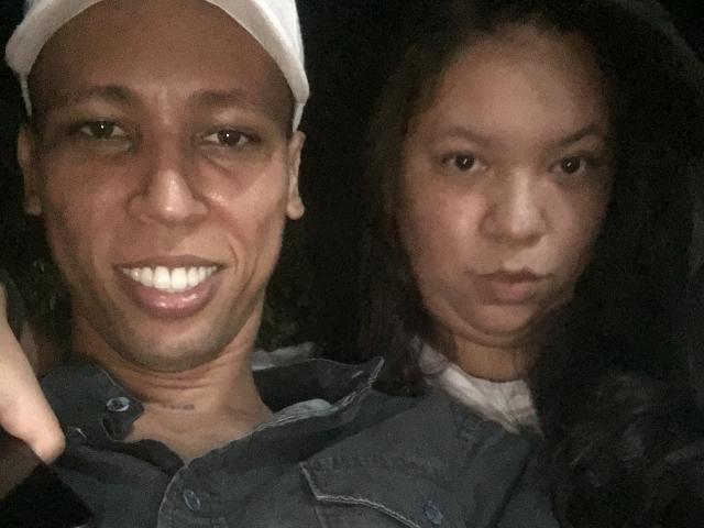 ElChicletv with tan skin, white cap, and smiling teeth, next to ElChicletv with darker skin, long hair, and serious expression, both wearing denim shirts, in dim light.