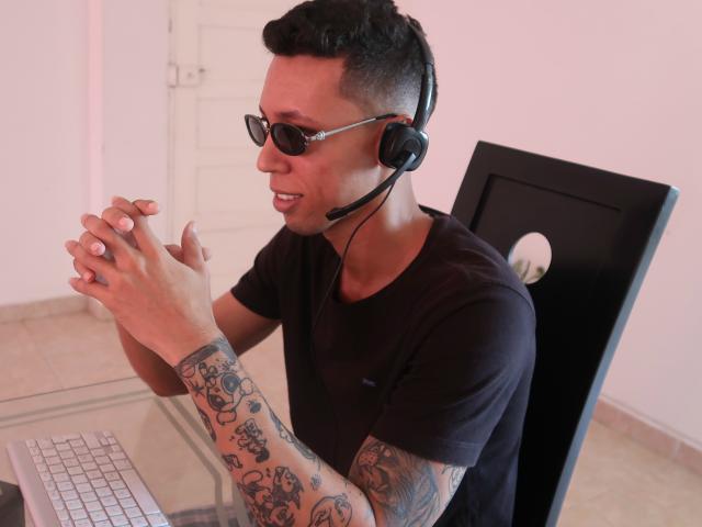 ElChicletv, wearing black t-shirt, black headphones, sunglasses, and tattoos on arms, typing on white keyboard, hands raised, smiling, black chair, short black hair, looking to the side.